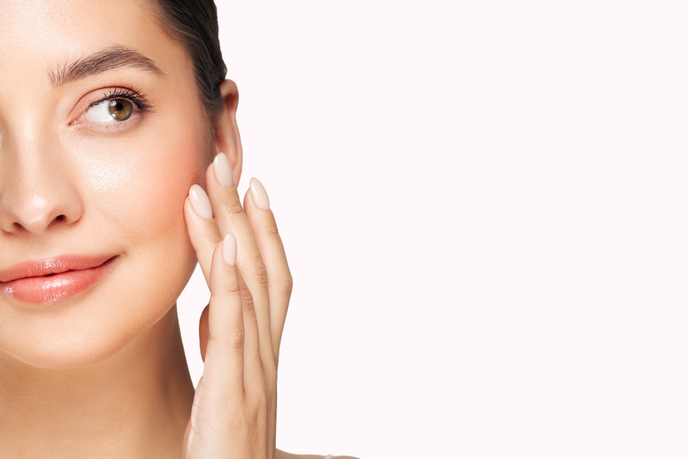 A close-up shot of a woman with clear skin smiling and touching her cheek, isolated against a white background.