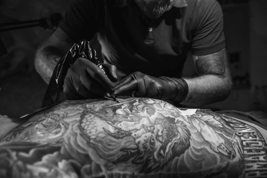 A black and white image of a tattoo artist working on a large, detailed chest tattoo.