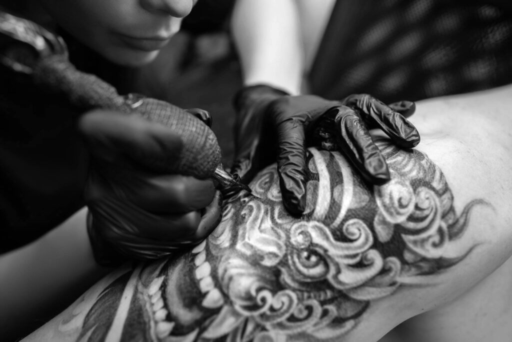 A close-up, black and white image of a tattoo artist in black gloves tattooing an intricate Japanese-style design onto a client's leg or knee.