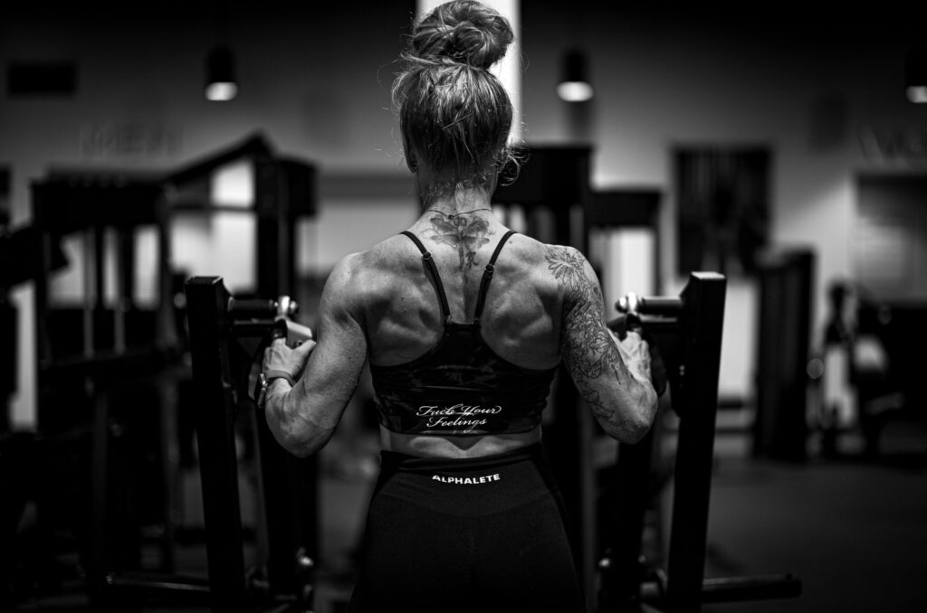 A black and white image from behind of a muscular woman with a bun and back tattoos using a row machine in a gym.