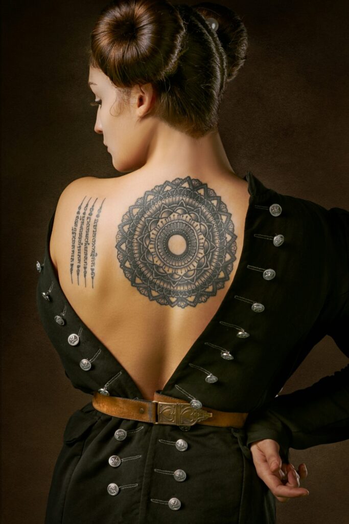 A rear view of a woman with her hair in twin buns, wearing a low-backed black jacket that displays a large mandala tattoo on her upper back and a script tattoo on her shoulder blade.