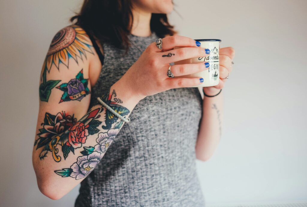 A cropped shot of a woman with colourful traditional-style sleeve tattoos, wearing rings and blue nail polish while holding a campfire-style mug.