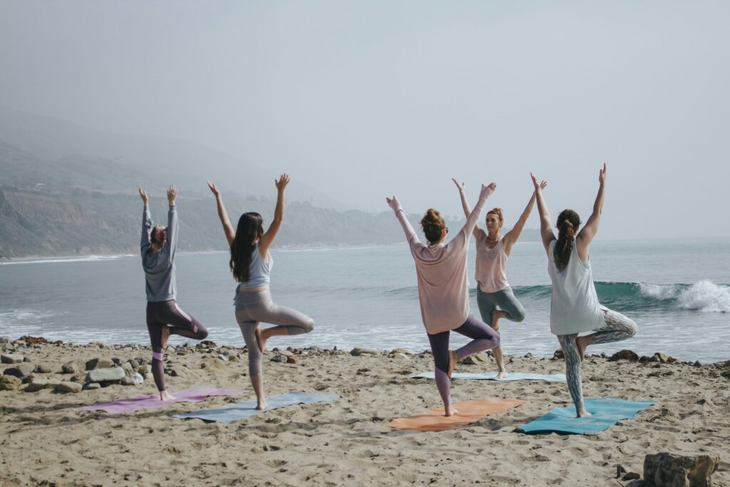 A group of women seen from behind practicing the tree yoga pose on mats at a sandy beach overlooking the ocean on a hazy day.