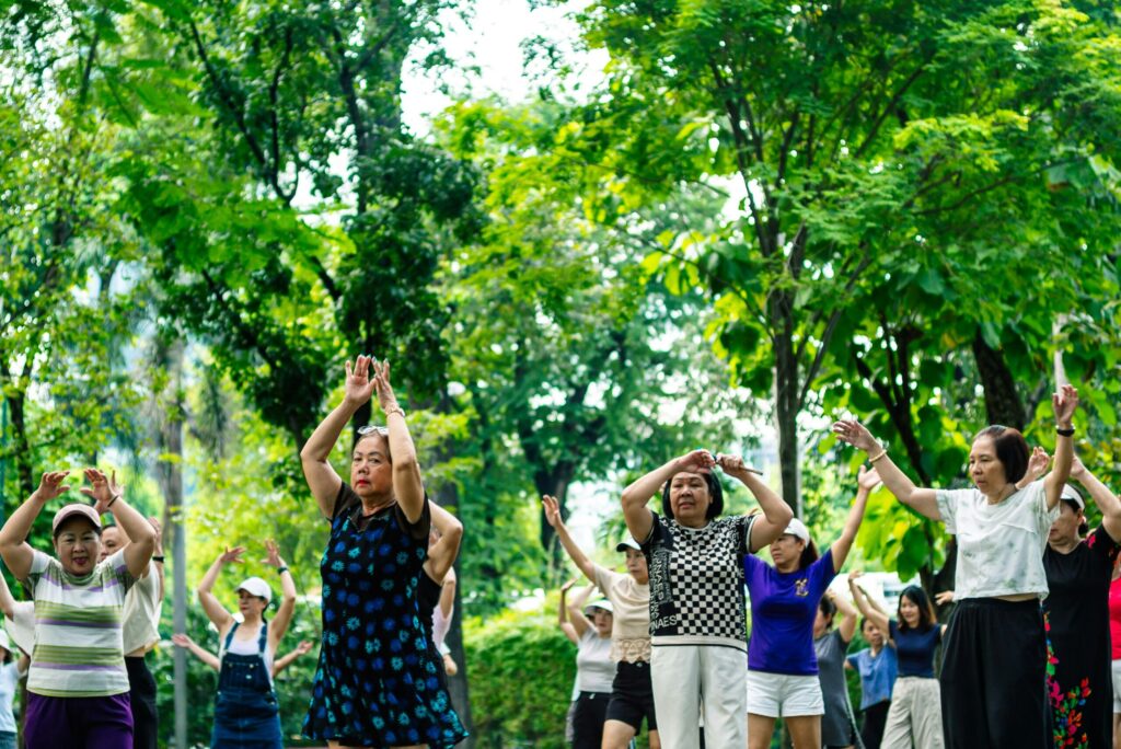 A large group of elderly women practicing tai chi or aerobics together in a lush, tree-filled public park.