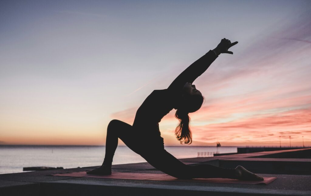 A silhouette of a woman performing a crescent lunge yoga pose on a mat outdoors, set against a colorful sunrise over the water.