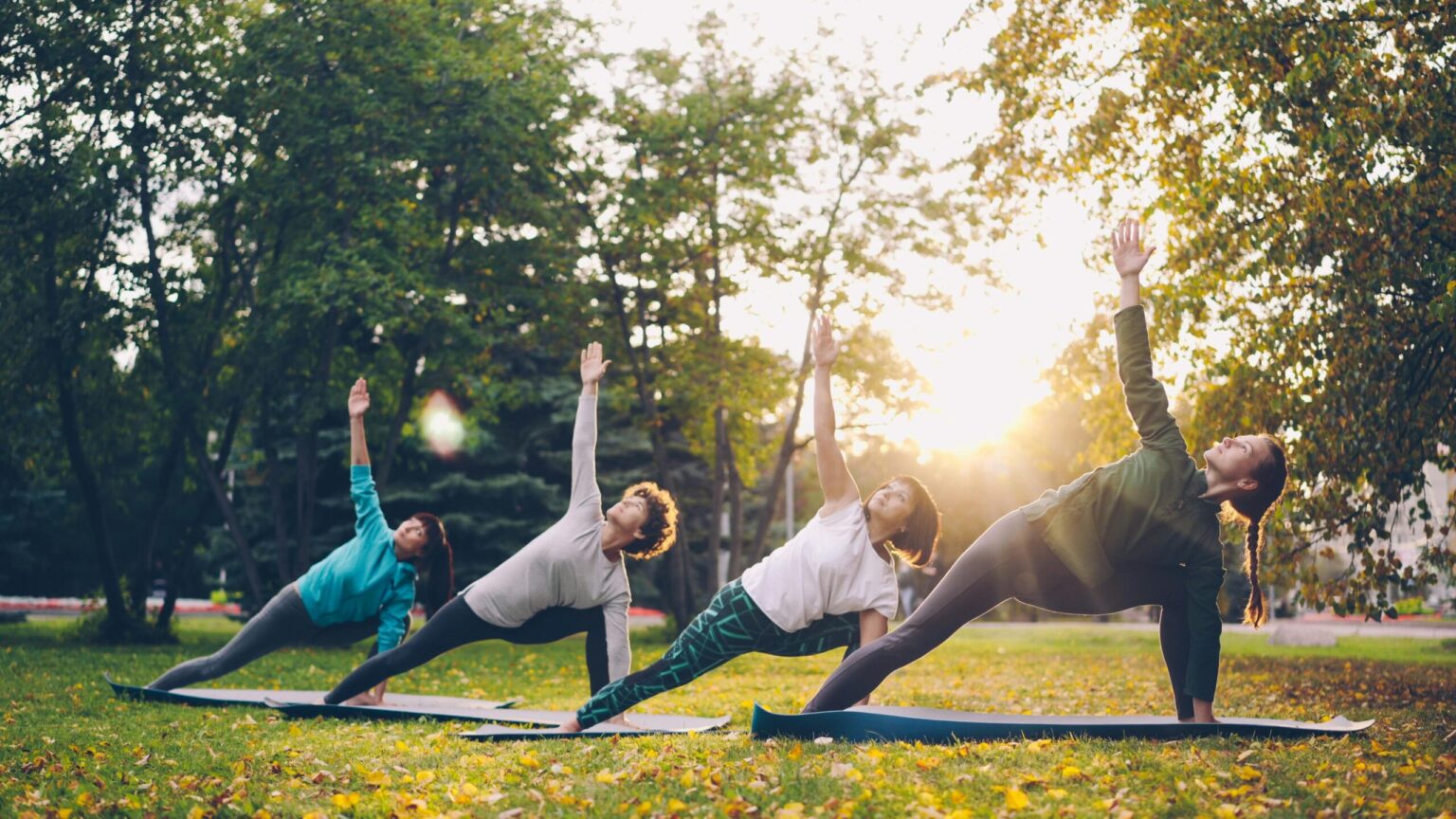 A group of four women practicing side angle yoga poses on mats in a grassy park during a bright sunset.