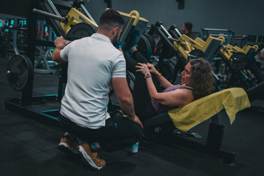 A personal trainer in a white shirt spots a woman as she uses a leg press machine in a gym.