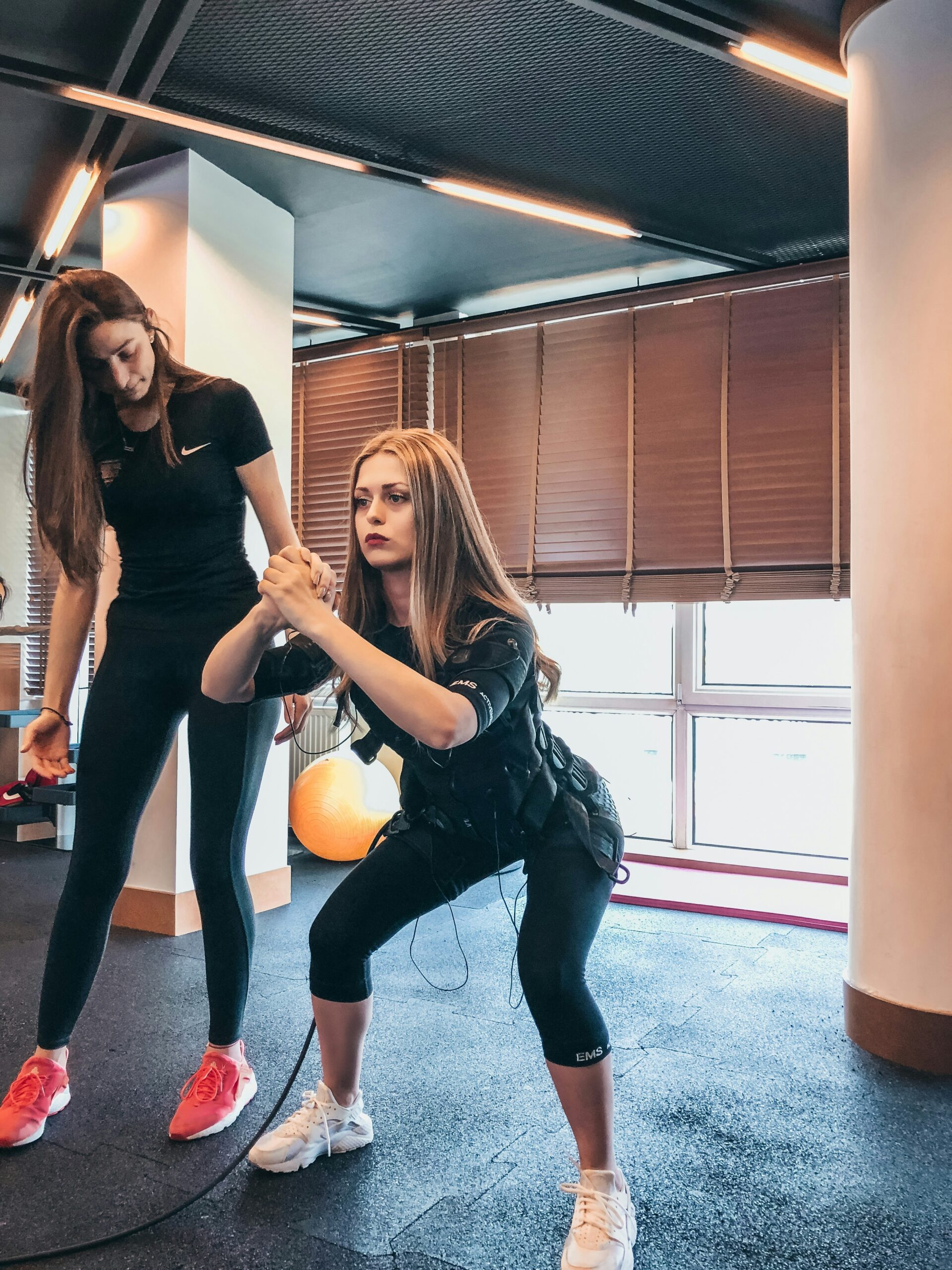 A woman wearing an EMS (Electrical Muscle Stimulation) training suit performing a squat while a personal trainer guides her in a gym.