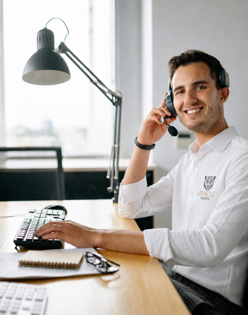 A smiling man wearing a headset and a white shirt with a "Muscle Tattoo" logo sits at a wooden desk.