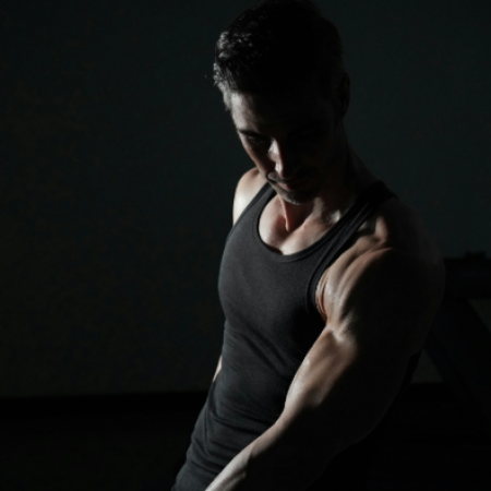 A athletic man in a black tank top looking down, highlighting a muscular arm under dramatic lighting against a dark background.