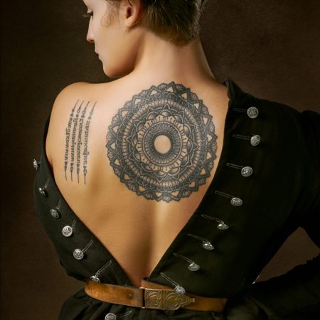 A rear view of a woman with her hair in twin buns, wearing a low-backed black jacket that displays a large mandala tattoo on her upper back and a script tattoo on her shoulder blade.