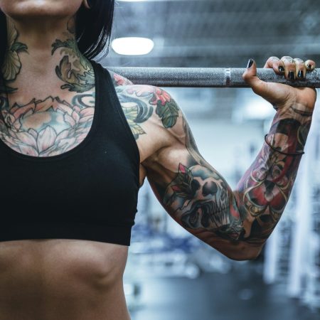 A cropped shot of a muscular woman with extensive chest, neck, and arm tattoos, holding a barbell on her shoulders inside a gym.