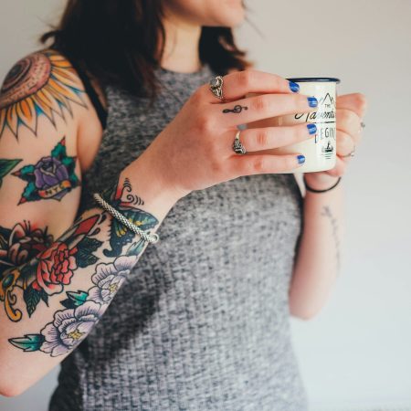 A cropped shot of a woman with colourful traditional-style sleeve tattoos, wearing rings and blue nail polish while holding a campfire-style mug.