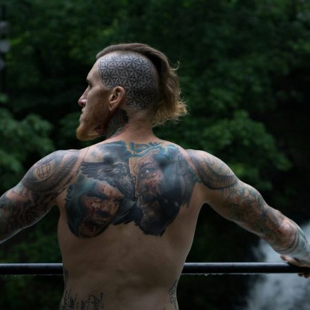 A back view of a shirtless man with extensive head, back, and arm tattoos, overlooking a waterfall.