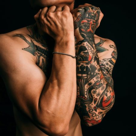 A cropped shot of a shirtless man with his arms crossed over his chest, showcasing an American traditional-style sleeve tattoo on his left forearm against a black background.