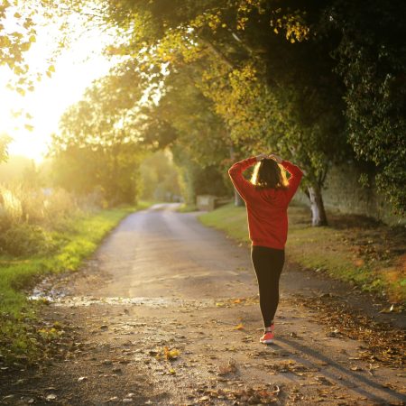 A rear view of a woman in a red hoodie and leggings walking down a leaf-strewn country road during a bright sunset.
