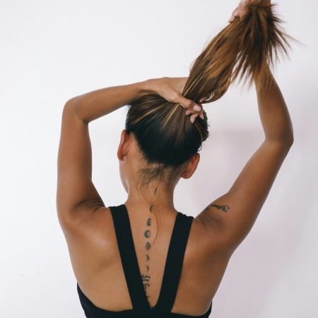 A rear view of a woman in a black sports bra tying her hair up, displaying a moon phase tattoo running down her spine against a plain white background.