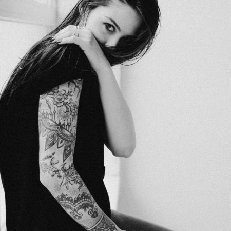 A black and white image of a woman partially obscuring her face with her arm, showcasing a detailed floral and mandala-style sleeve tattoo.