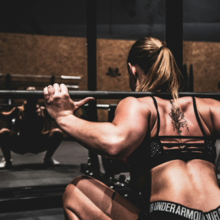 A rear view of a muscular woman with a spine tattoo performing a barbell squat in front of a gym mirror.