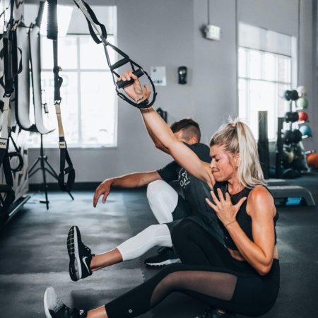 A man and a woman performing pistol squats together in a gym while holding onto suspension straps for support.