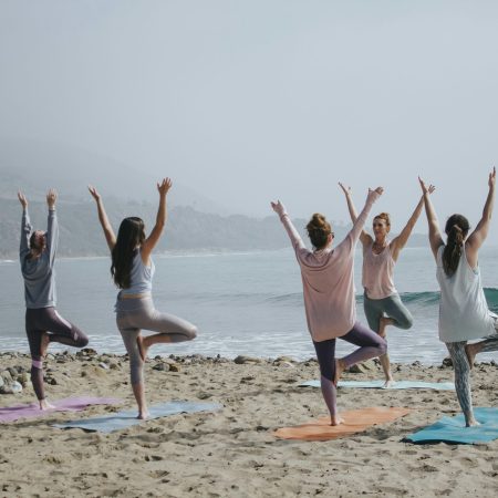 A group of women seen from behind practicing the tree yoga pose on mats at a sandy beach overlooking the ocean on a hazy day.