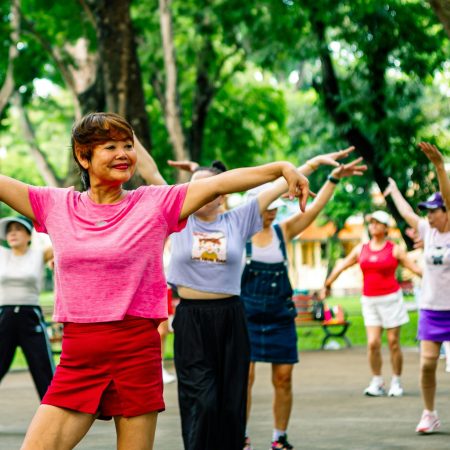 A woman in a pink shirt and red shorts leads an outdoor aerobics or dance class for a group of women in a green park.