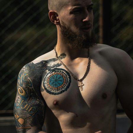 A shirtless, bearded man with a shaved head displaying a Norse-themed chest and sleeve tattoo while wearing a gold cross necklace.