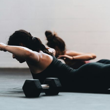 Two women lying face down on a gym floor while performing back extensions or a superman exercise, with a dumbbell in the foreground.