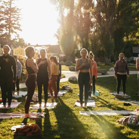 A group of women stand on yoga mats in a grassy park, placing their hands over their hearts during an outdoor class at sunset.