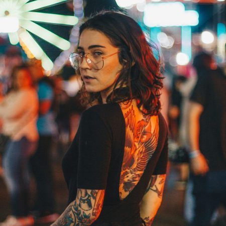 A young woman with glasses looking back over her shoulder, showing a large tattoo on her open-backed black shirt, with a blurred carnival and Ferris wheel in the background.