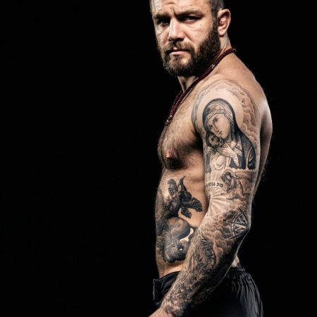 A side profile of a shirtless, bearded man showcasing an intricate sleeve tattoo featuring religious imagery against a black background.