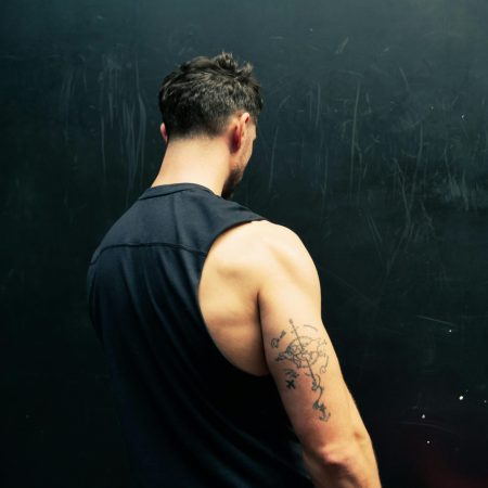 A man seen from behind wearing a black sleeveless shirt, showcasing a compass and world map tattoo on his upper arm against a dark background.