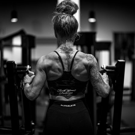 A black and white image from behind of a muscular woman with a bun and back tattoos using a row machine in a gym.